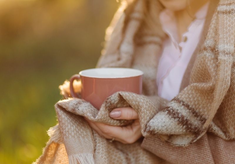 xícara na mão de uma jovem, coberta com um cobertor num pôr do sol
