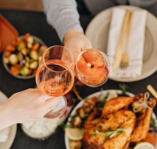 Mãos de duas pessoas brindando, com uma mesa em desfoque com alimentos de ceia de natal.